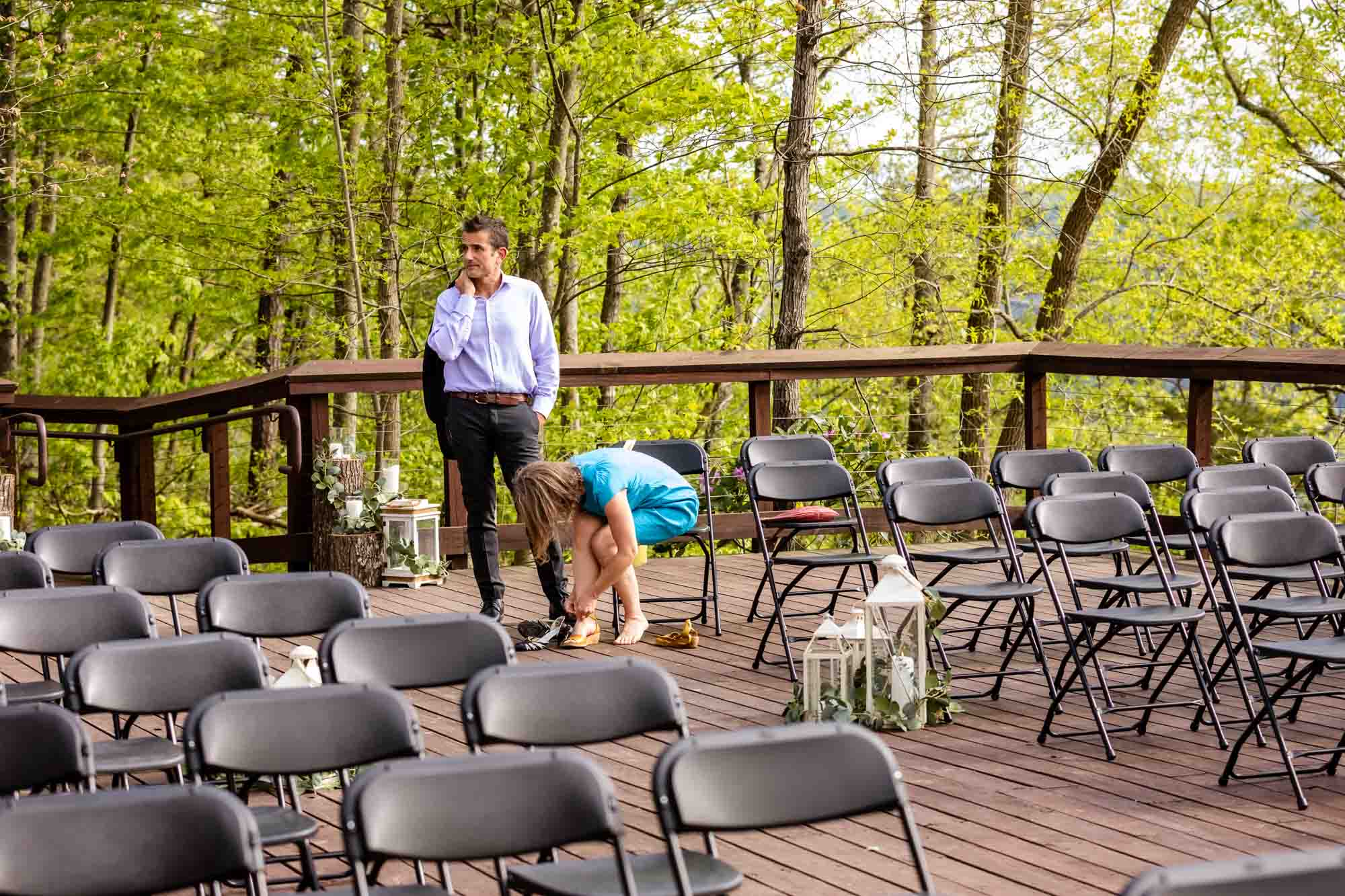 funny moment at wedding ceremony while man waits for his partner as she fixes her shoes, surrounded by empty chairs, with his suit coat thrown over his shoulder and an impatient look on his face.
